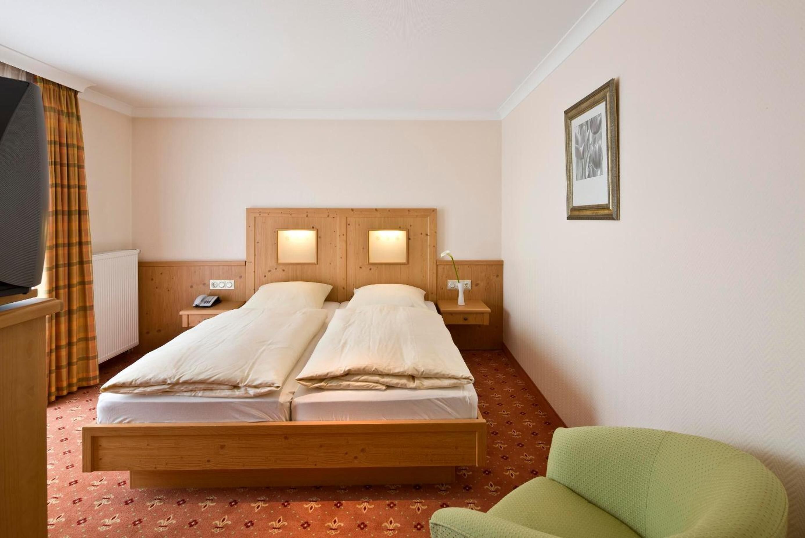 A double bed with wooden headboard and nightstands, a green armchair, and a framed picture on the wall.