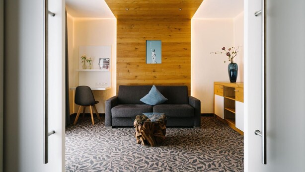A modern living room with a wooden ceiling, a grey sofa with a blue pillow, a glass coffee table, and a wooden sideboard with a vase. - Germanenhof (Steinheim)