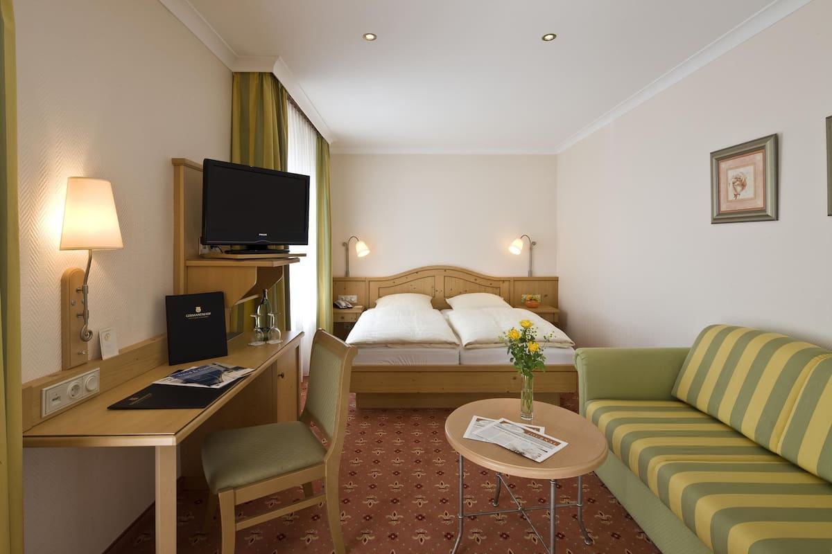 A hotel room with a bed, a TV, a desk with a lamp, a chair, a sofa, and a small table with a vase of flowers.