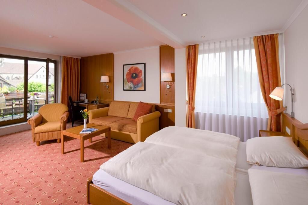 A hotel room with a bed, a sofa, a chair, a coffee table, and a desk. There is a window with curtains, a painting on the wall, and a view of buildings outside.