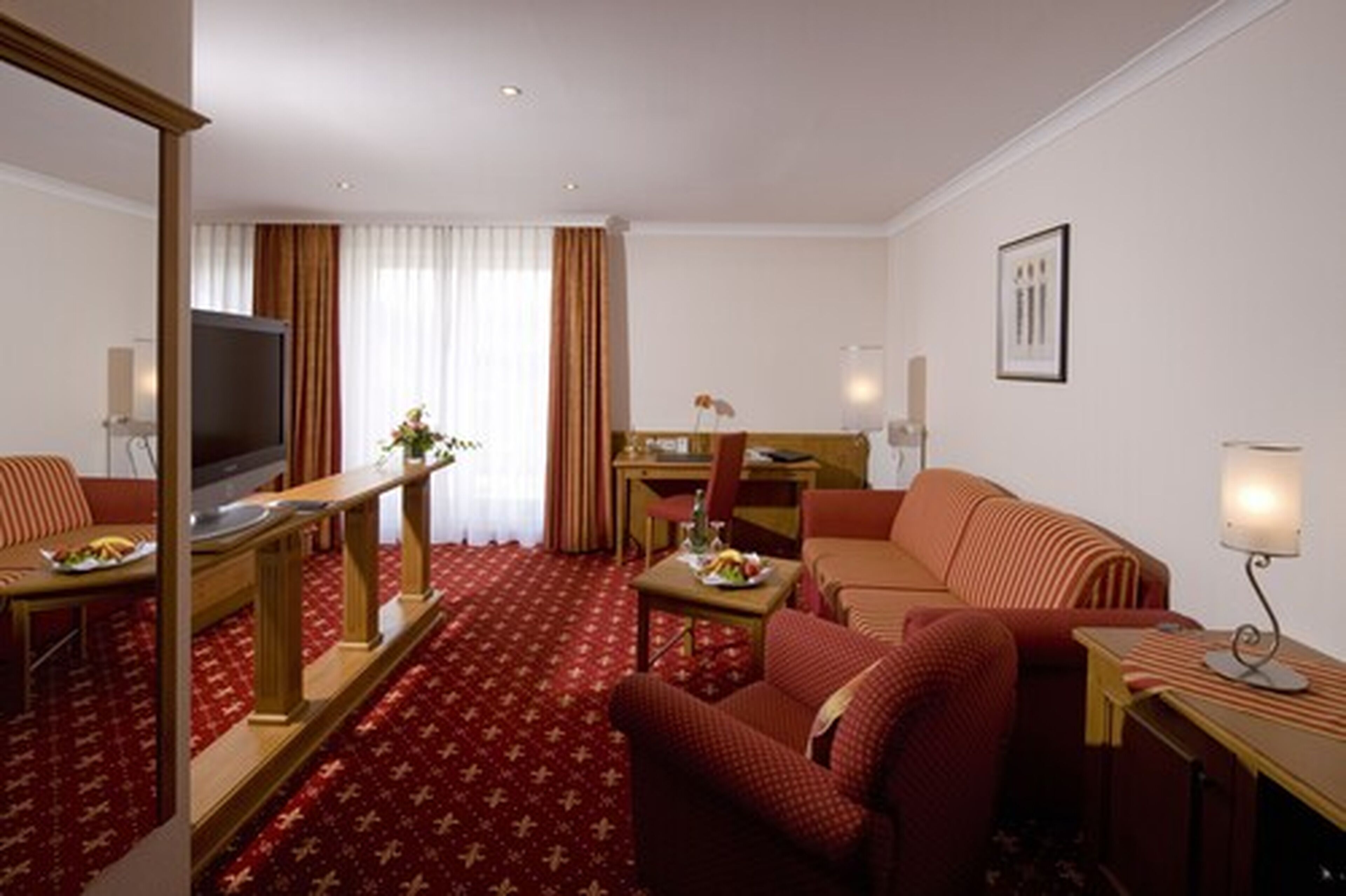 A hotel room with a red carpet, a television, a dining table with chairs, a sofa, and a desk with a lamp.