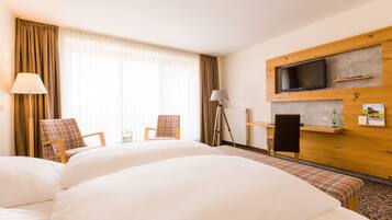 A hotel room with two beds, a TV mounted on a wooden wall, a desk with a lamp, and a window with curtains.