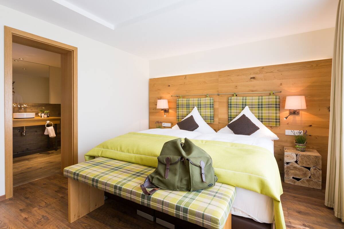 A hotel room with a wooden headboard, a bed with green bedding, a bench with a backpack, and a small bedside table with a lamp.