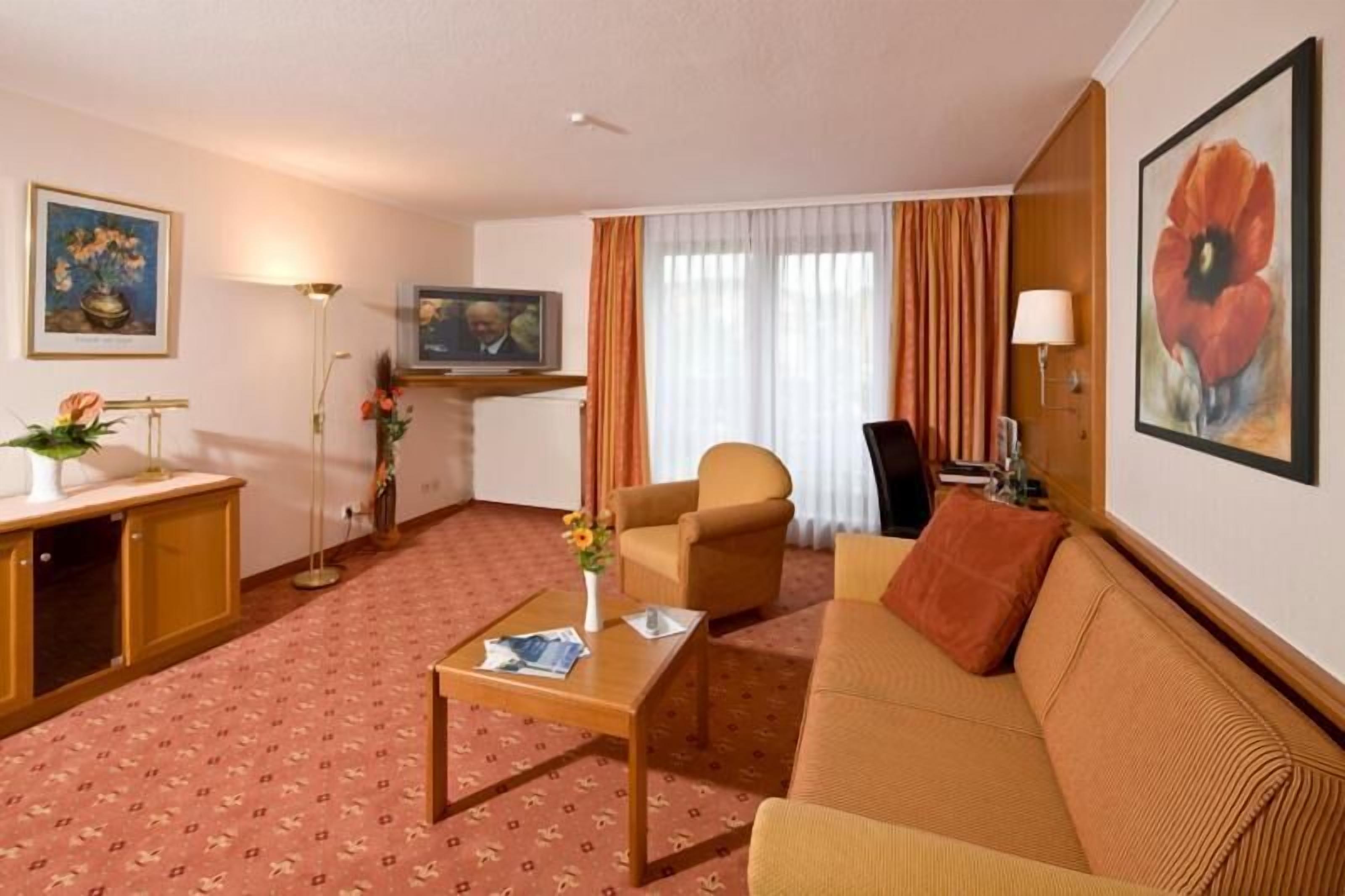 Quarto de hotel com sofá, poltrona, mesa de centro, televisão e obras de arte nas paredes.