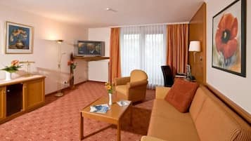 Quarto de hotel com sofá, poltrona, mesa de centro, televisão e obras de arte nas paredes.