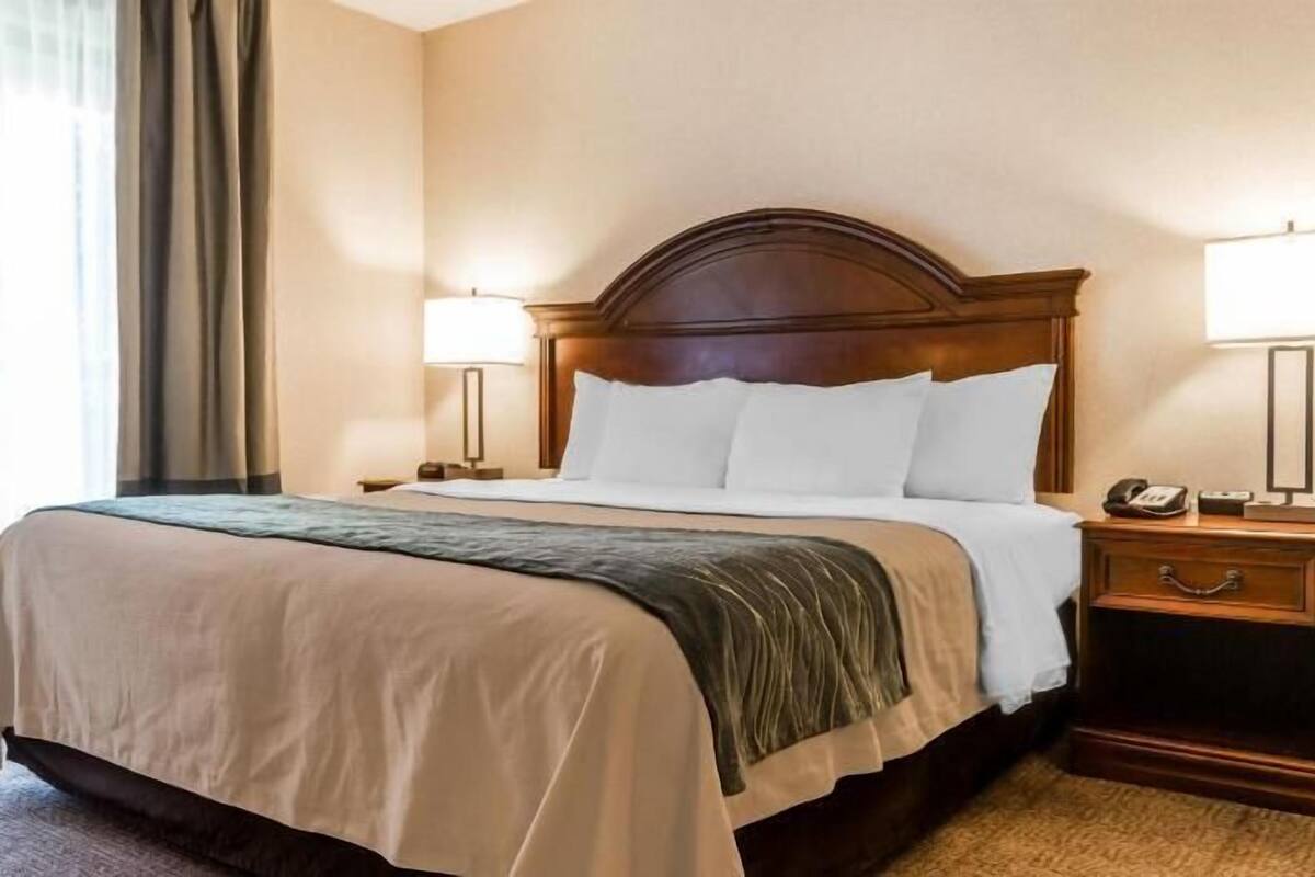 Ein Hotelzimmer mit einem großen Bett, zwei Nachttischen, einem Sessel und einem fenster mit Vorhängen.