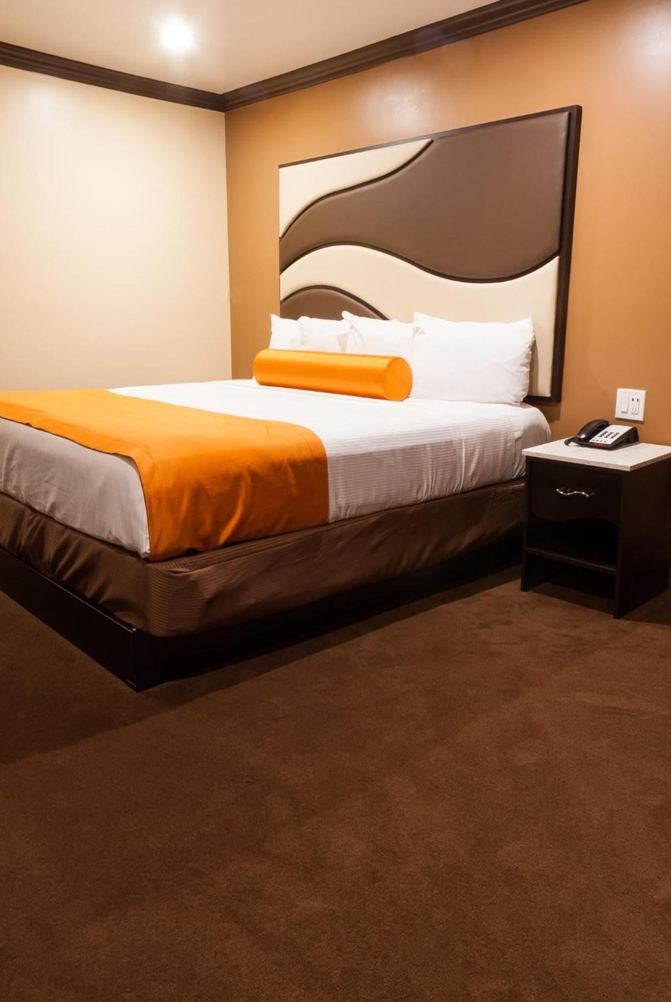 A hotel room with a large bed, an orange throw blanket, a nightstand with a phone, and a wall-mounted light.