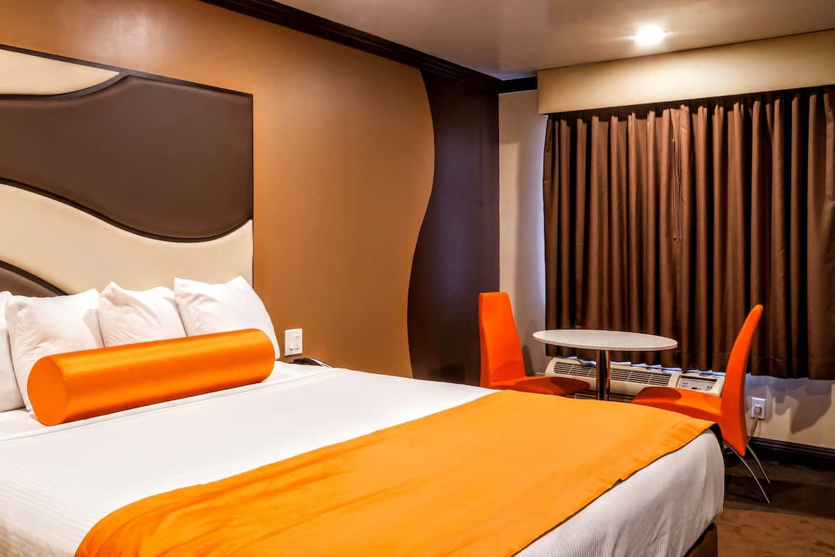 A hotel room with a large bed, white and orange bedding, a modern headboard, an orange chair, a small round table, and a window with curtains.