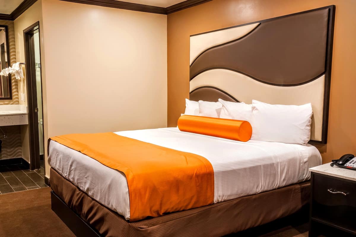A hotel room with a bed, an orange accent pillow, a white comforter, a nightstand with a phone, and a bathroom with a sink and mirror.
