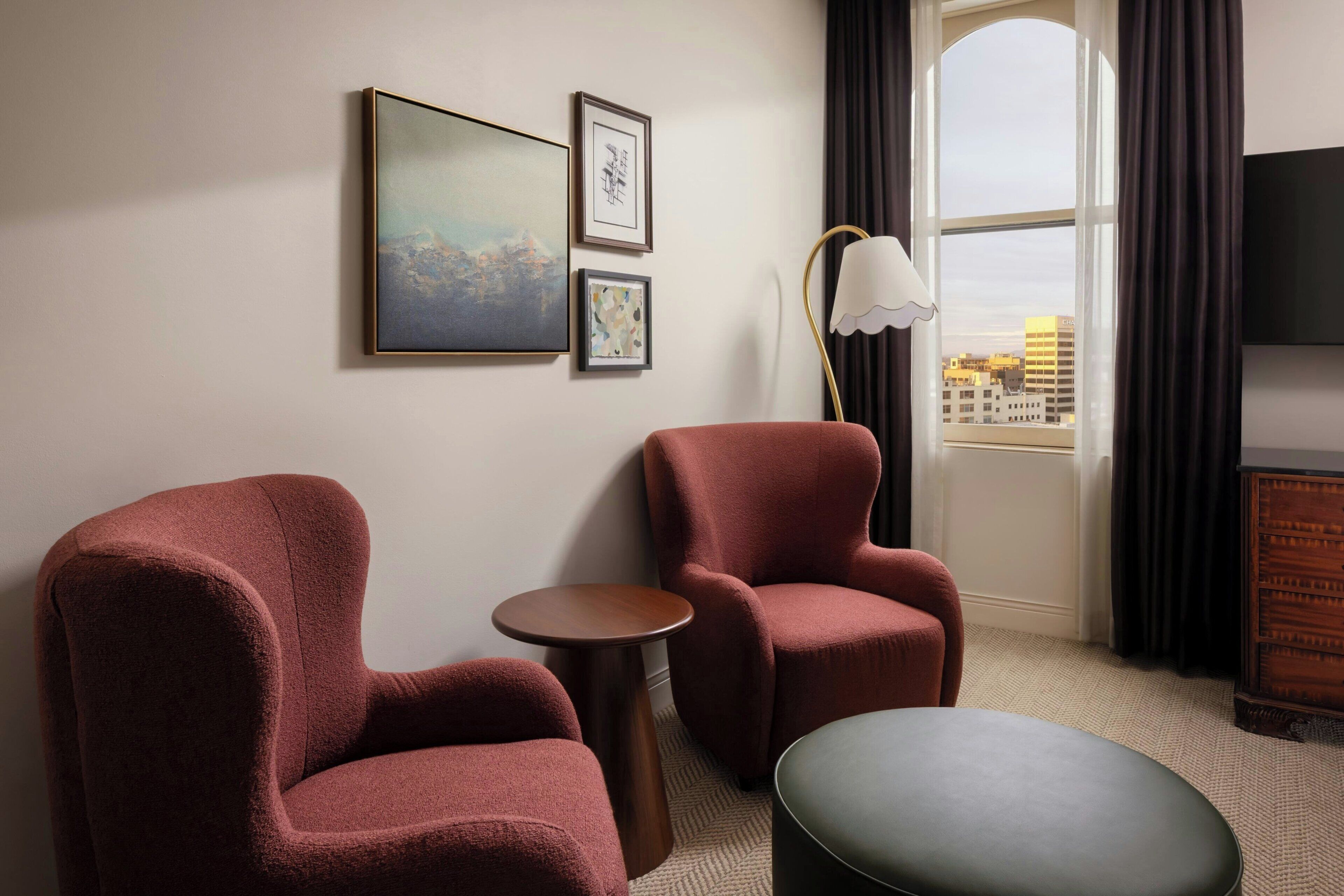 A hotel room with two armchairs, a small round table, a large window with curtains, and a flat-screen TV on a wooden cabinet.