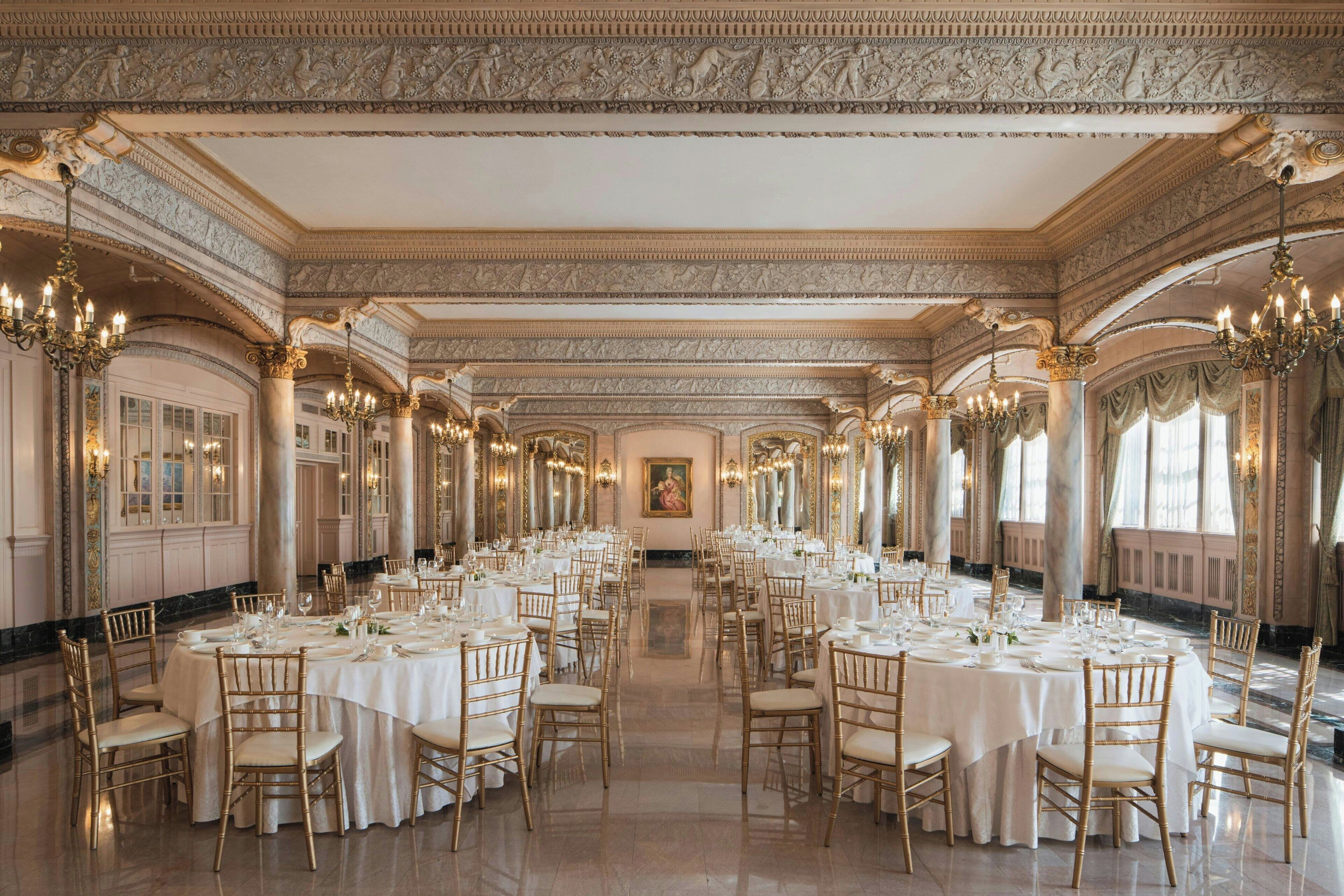 A grand banquet hall with ornate ceiling and columns, round tables set for dining, and chandeliers.