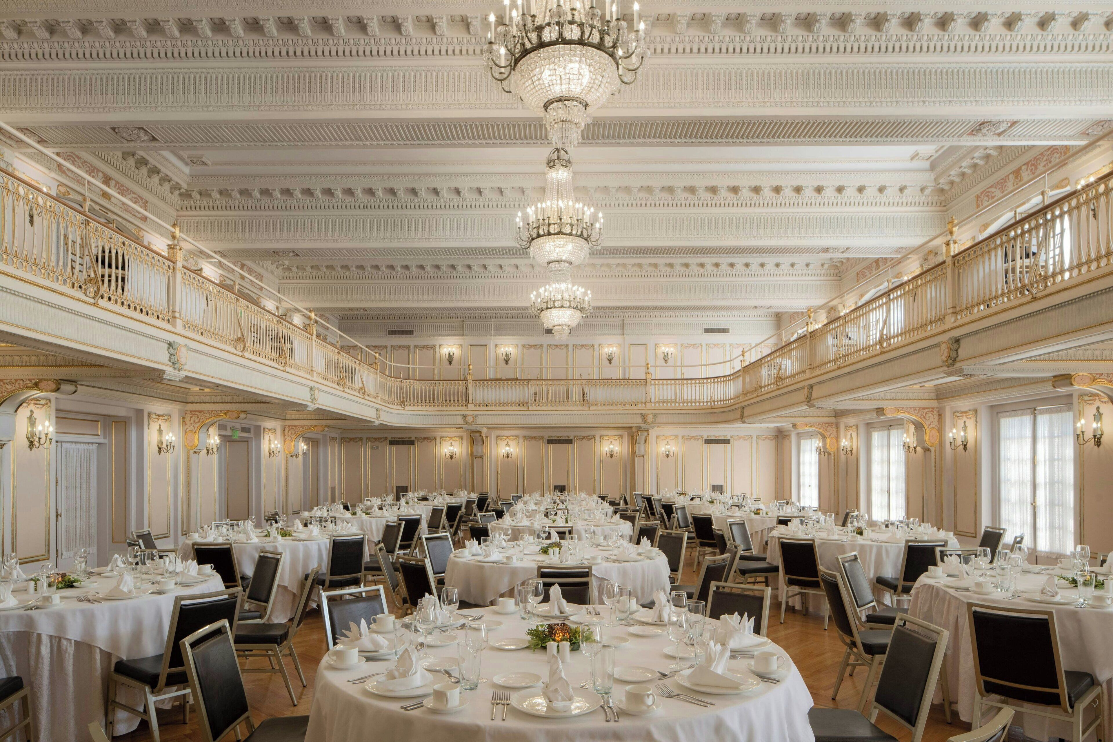 A grand banquet hall with ornate chandeliers, round tables set for dining, and a balcony overlooking the room.
