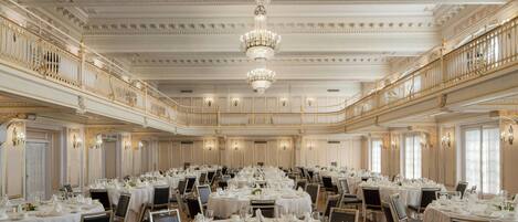 A grand banquet hall with ornate chandeliers, round tables set for dining, and a balcony overlooking the room.