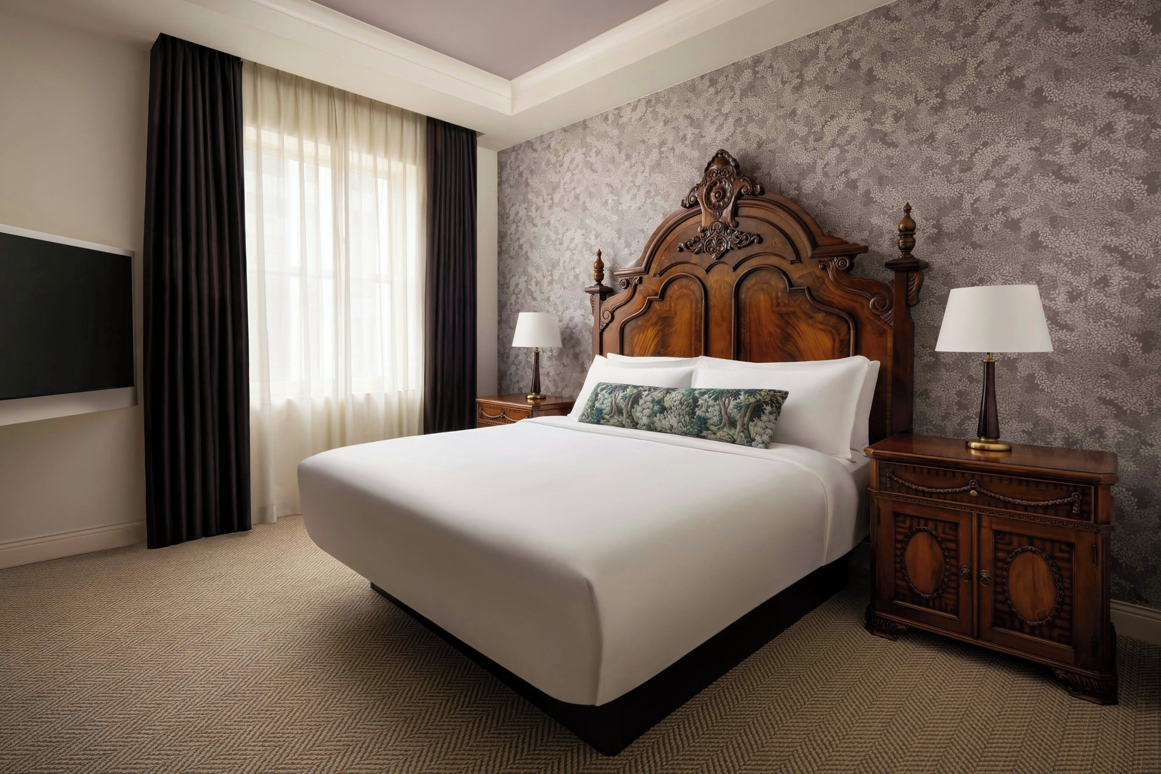 Habitación de hotel con una cama grande, dos mesitas de noche con lámparas, un televisor y una ventana con cortinas.