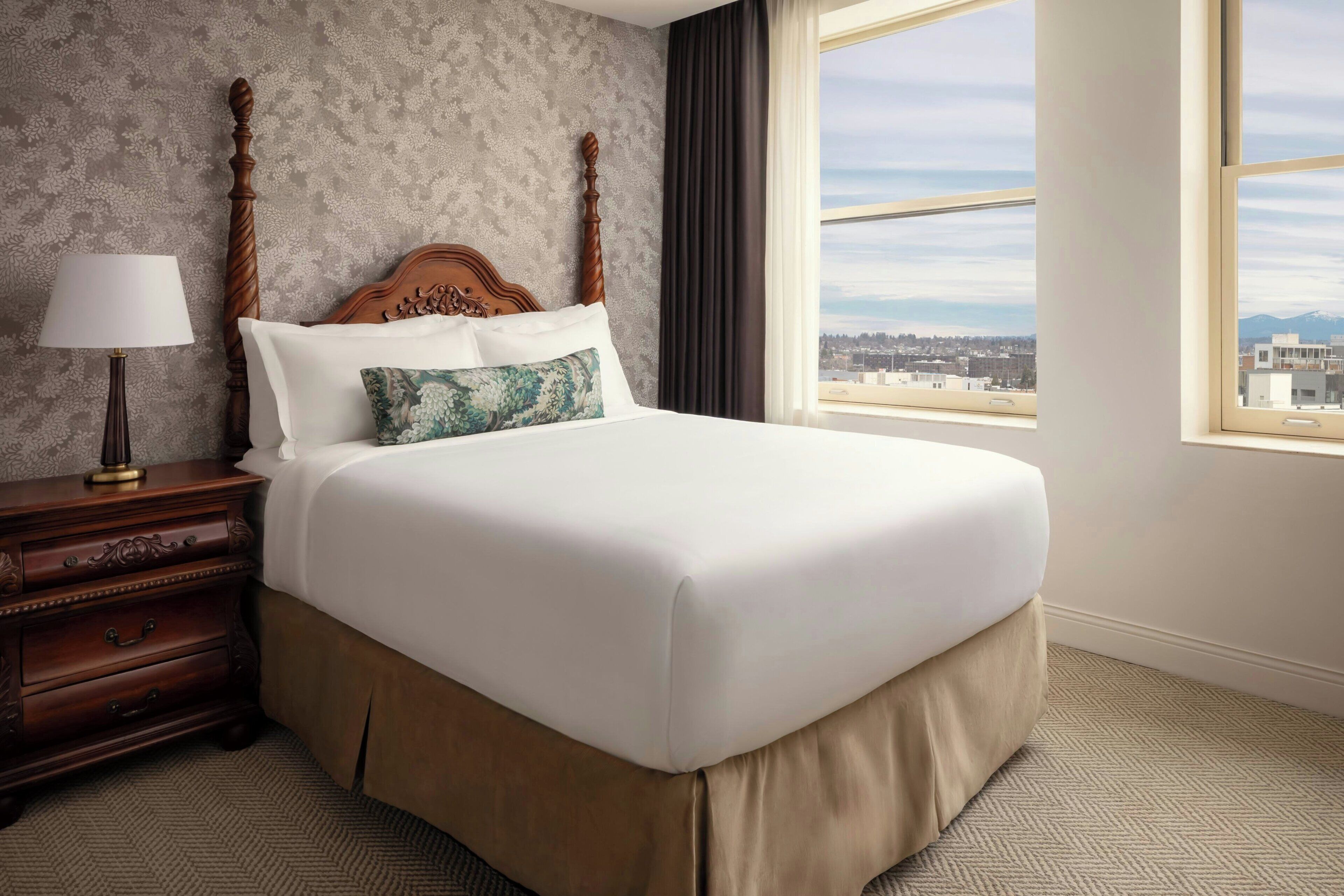 A neatly made bed with a wooden headboard, a nightstand with a lamp, and a view of the city through large windows.