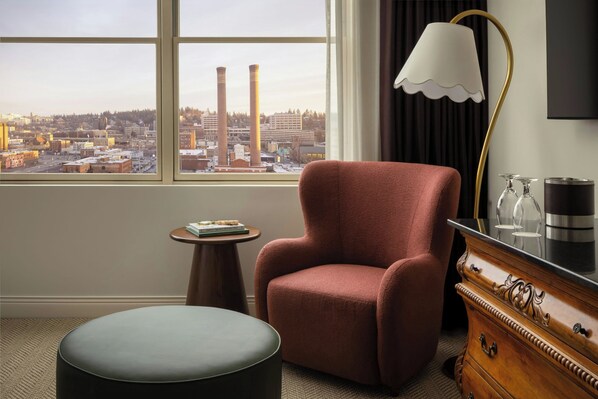 A room with a city view, a red armchair, a round ottoman, a small table with books, a lamp, and a dresser with a mirror.