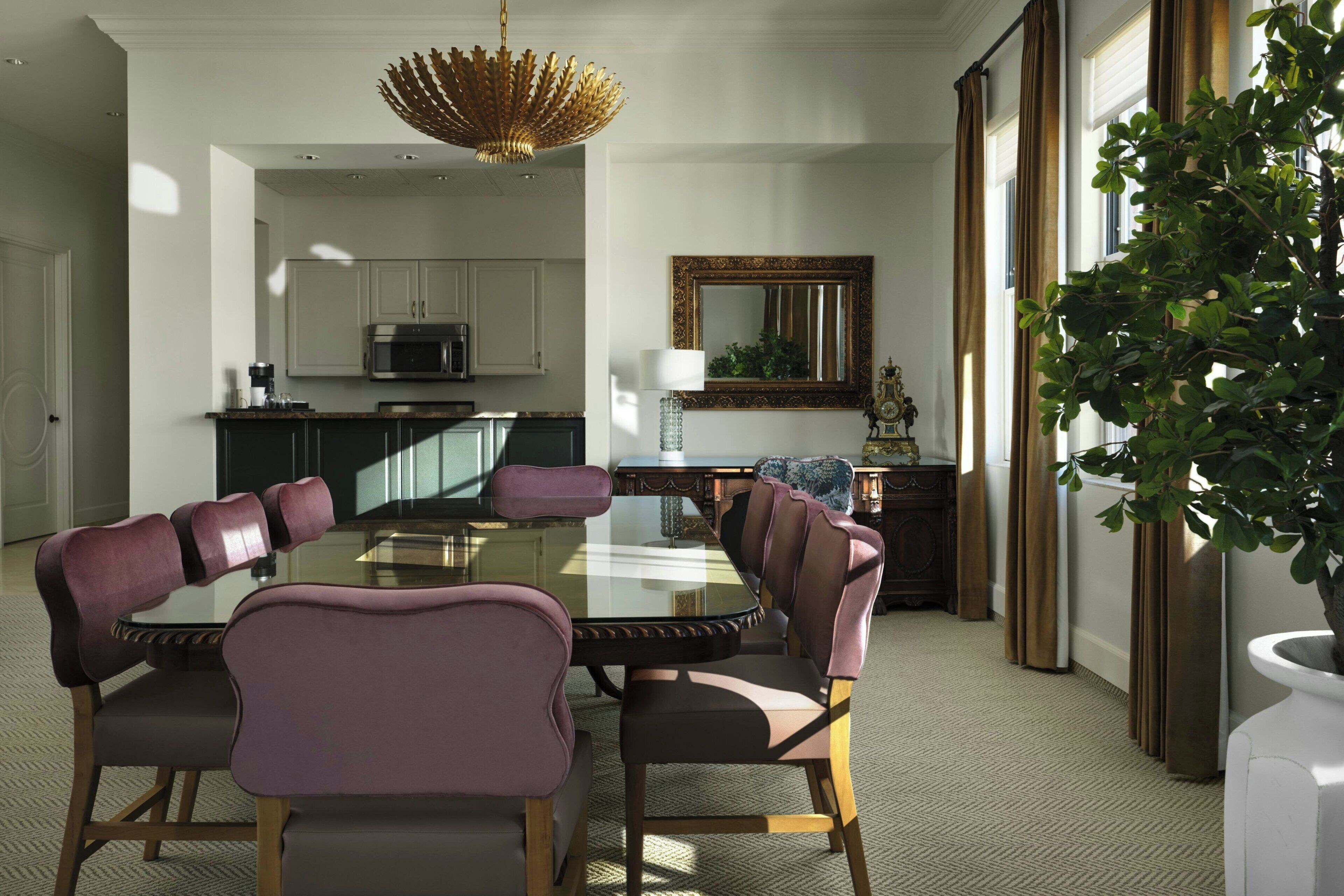Un'area pranzo con tavolo in vetro, sedie imbottite, un lampadario, una cucina sullo sfondo e un grande specchio a parete.