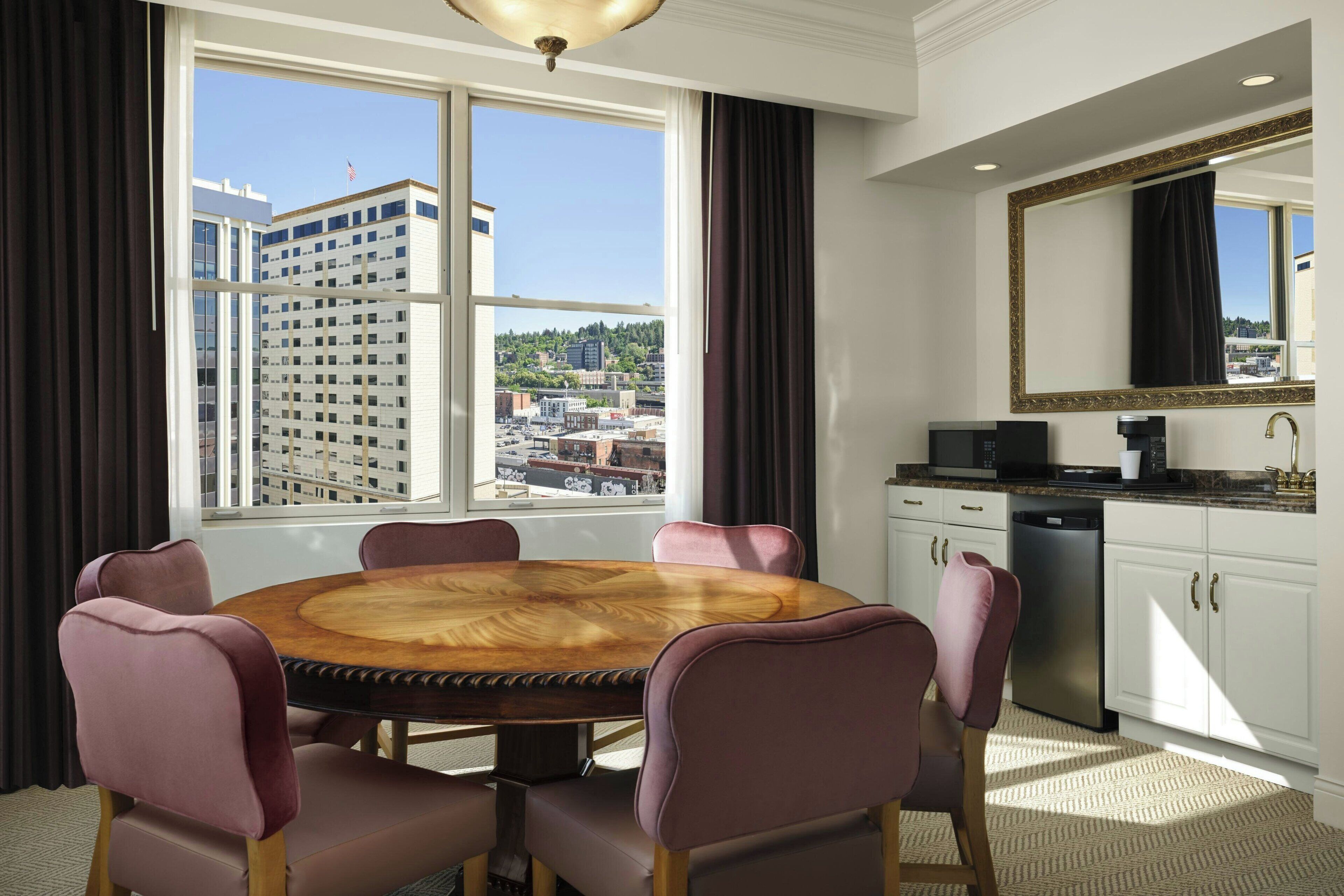 A hotel room with a round wooden dining table, six chairs, a kitchen area with a sink and microwave, and a large window offering a city view.