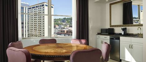 Habitación de hotel con una mesa de comedor redonda de madera, seis sillas, una zona de cocina con fregadero y microondas, y un amplio ventanal con vistas a la ciudad.