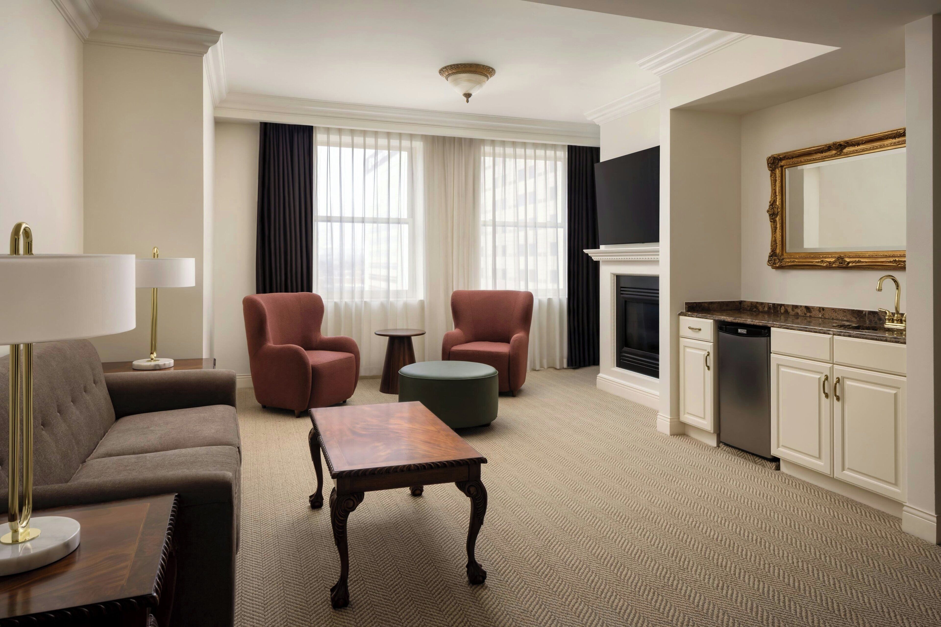 A hotel room with a sofa, two armchairs, a small table, a fireplace, a kitchen area with cabinets, and a large mirror.