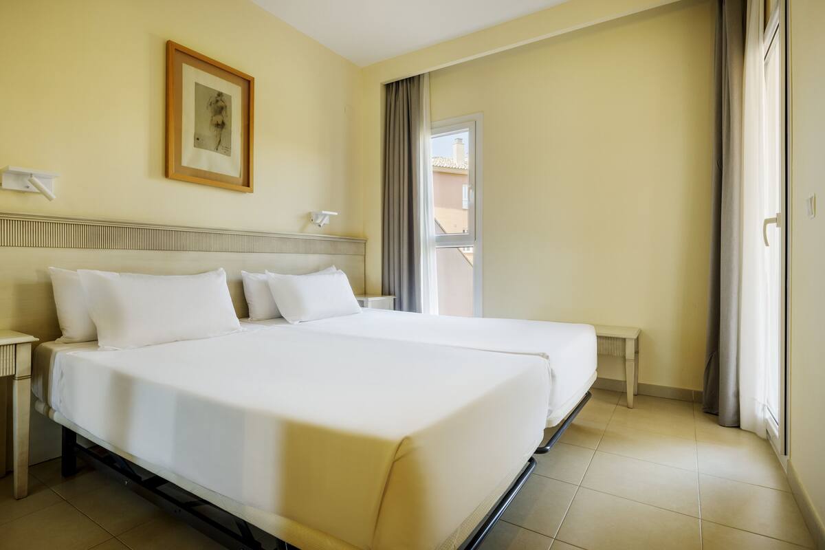 Une chambre d’hôtel avec un grand lit, une table de chevet, une petite table, un tableau encadré au mur et une fenêtre avec des rideaux.