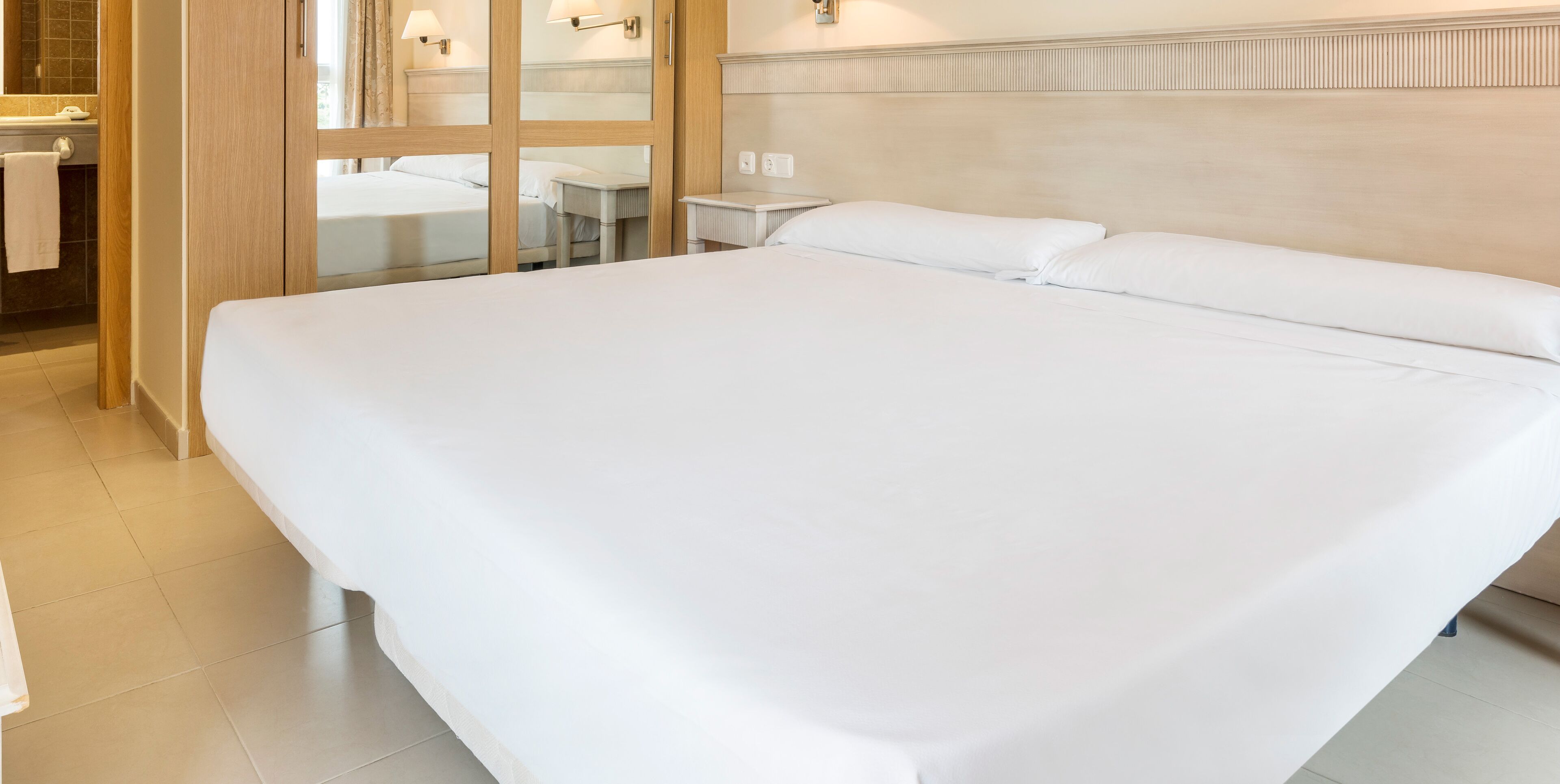 Una cama grande y bien hecha en una habitación de hotel, con cabecera de madera, una mesita de noche pequeña y un espejo.