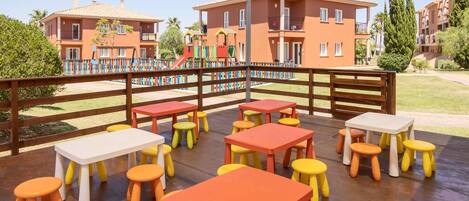 Un patio al aire libre con mesas y taburetes de colores, una cerca de madera y un parque infantil al fondo.