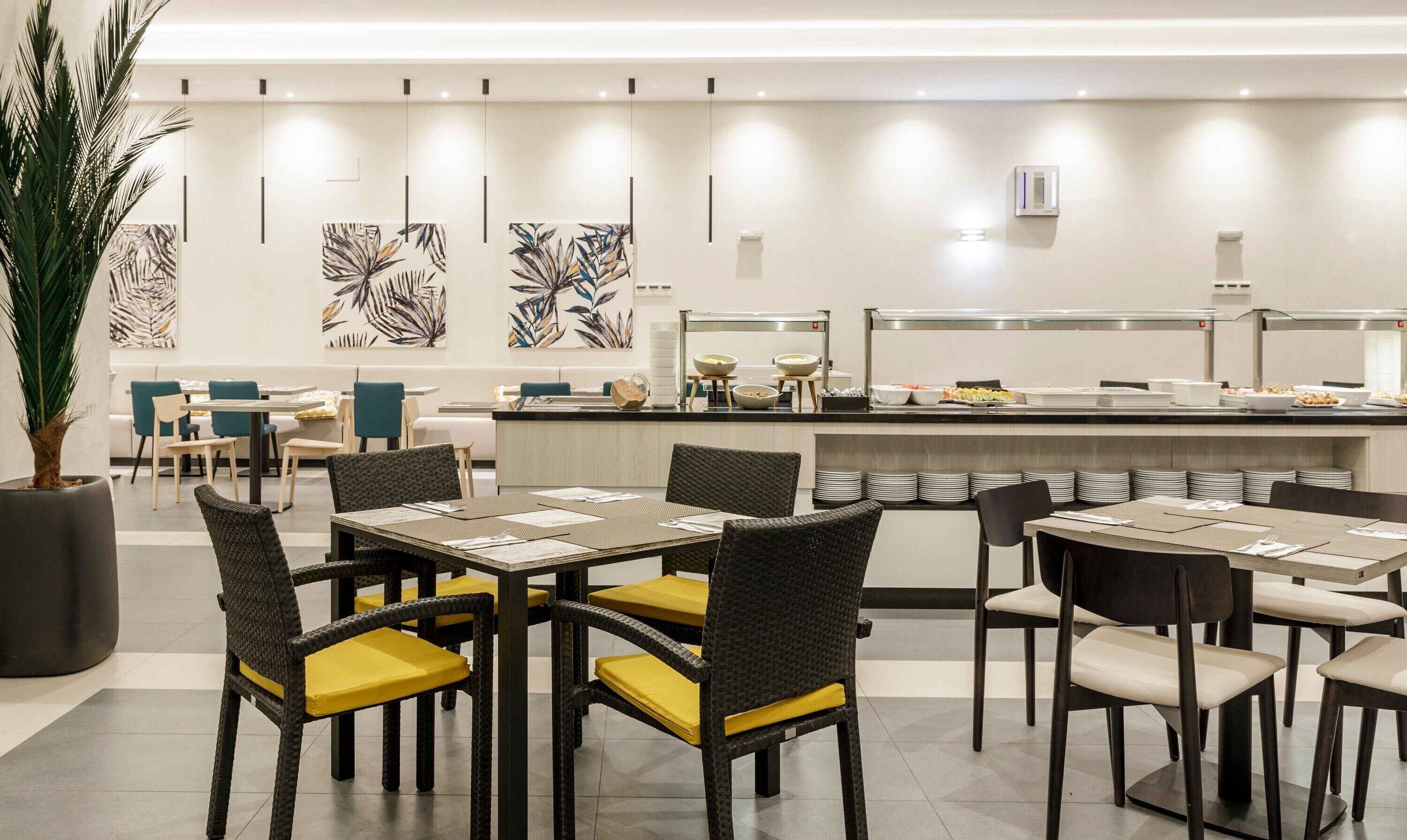 A modern dining area with wicker chairs and yellow cushions, a buffet counter with various food items, and abstract wall art.