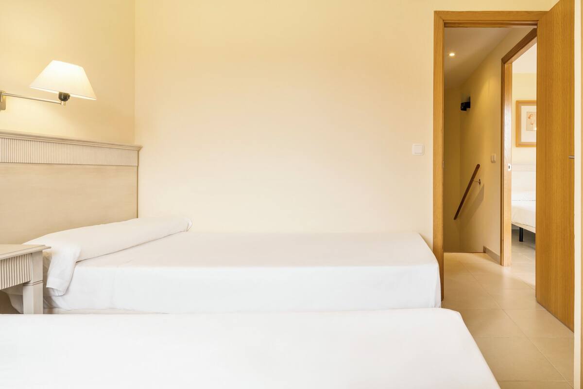 Une chambre d’hôtel avec deux lits, une lampe de chevet, une porte en bois et un couloir menant à une autre chambre.