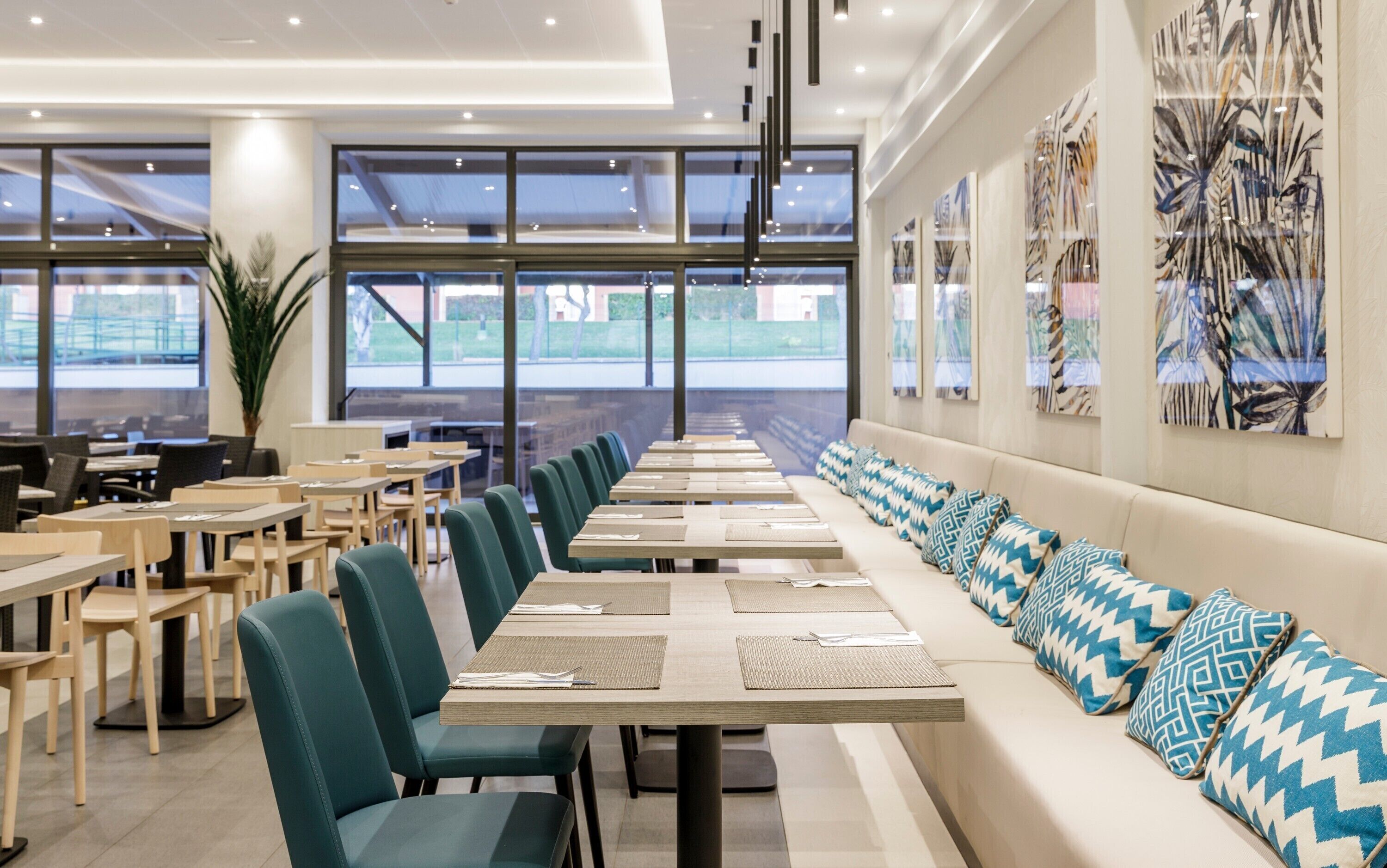 Un comedor moderno con mesas largas y sillas verdes, un sofá con cojines estampados y grandes ventanales que ofrecen vistas al exterior.