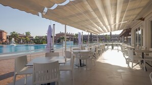 A poolside area with tables and chairs, a covered seating area, and a view of the pool and buildings in the background.