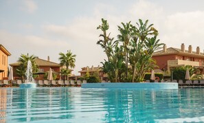 A swimming pool with a fountain, surrounded by lounge chairs and palm trees, with buildings in the background. - Aparthotel ILUNION Sancti Petri (Chiclana de la Frontera)