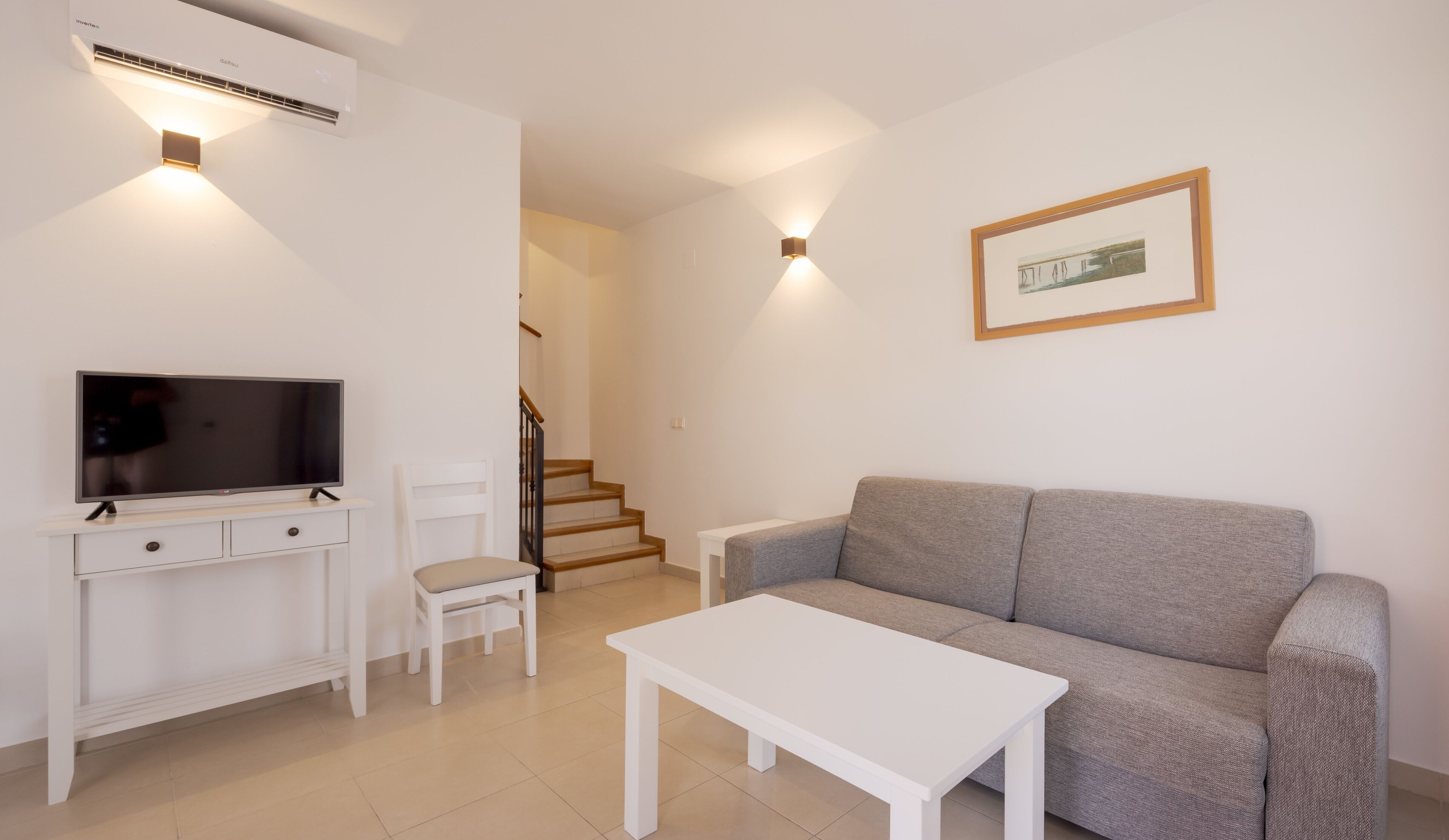 Una sala de estar con un sofá gris, una mesa de centro blanca, un televisor sobre un mueble blanco, una silla blanca y una escalera que lleva a otra habitación.