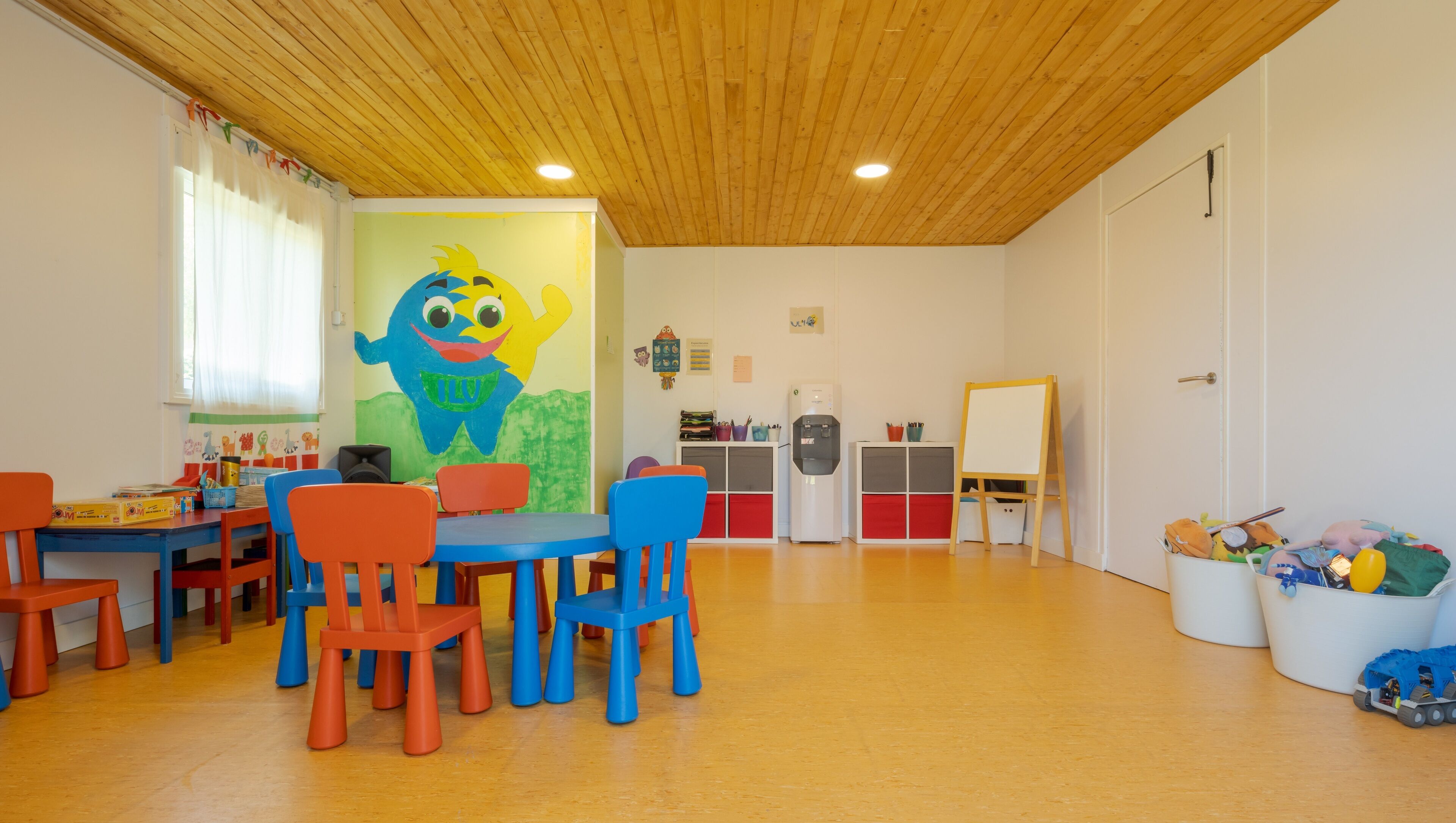 A children's playroom with wooden ceiling, colorful chairs, tables, and a large wall mural.