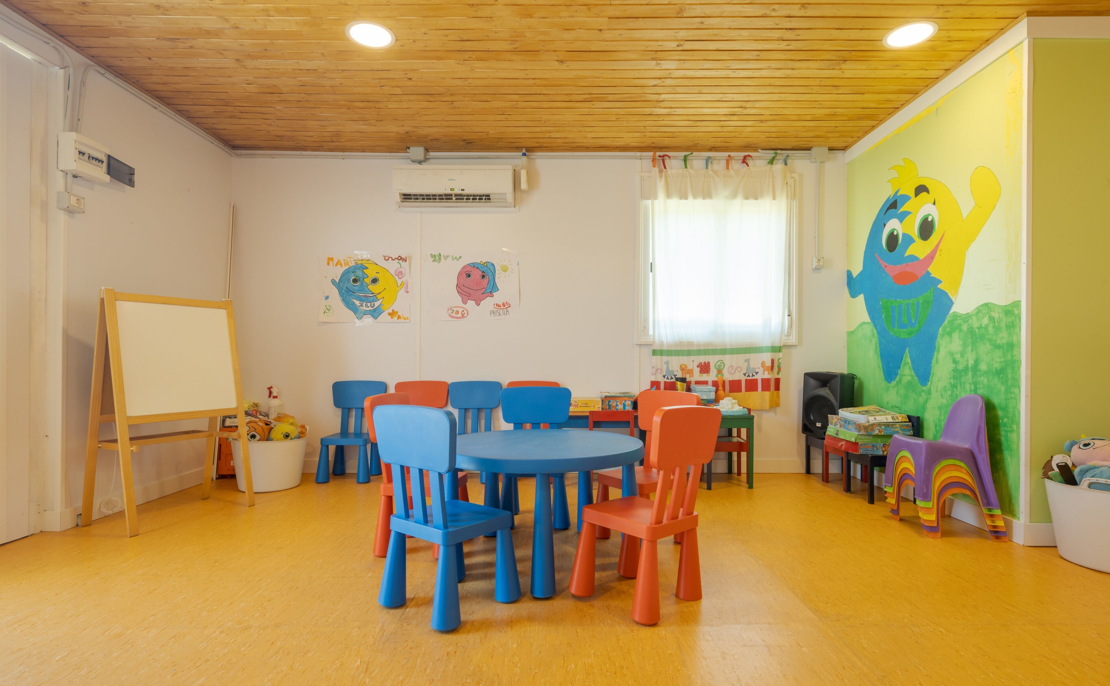 Una sala de juegos para niños con techo de madera, sillas coloridas, una pizarra y un gran mural de caricaturas en la pared.