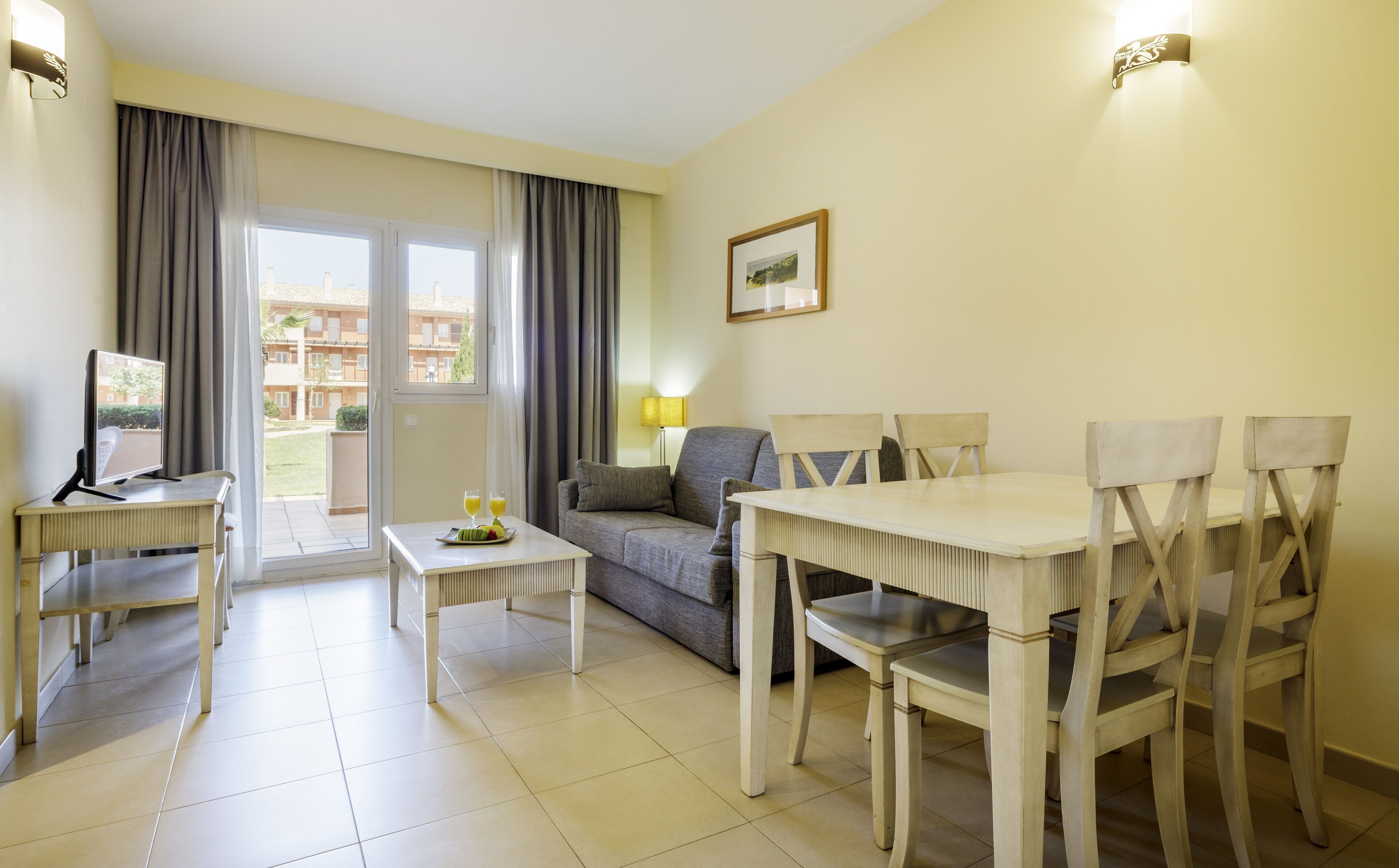 A furnished living room with a sofa, a dining table with chairs, a TV, and a window with curtains.