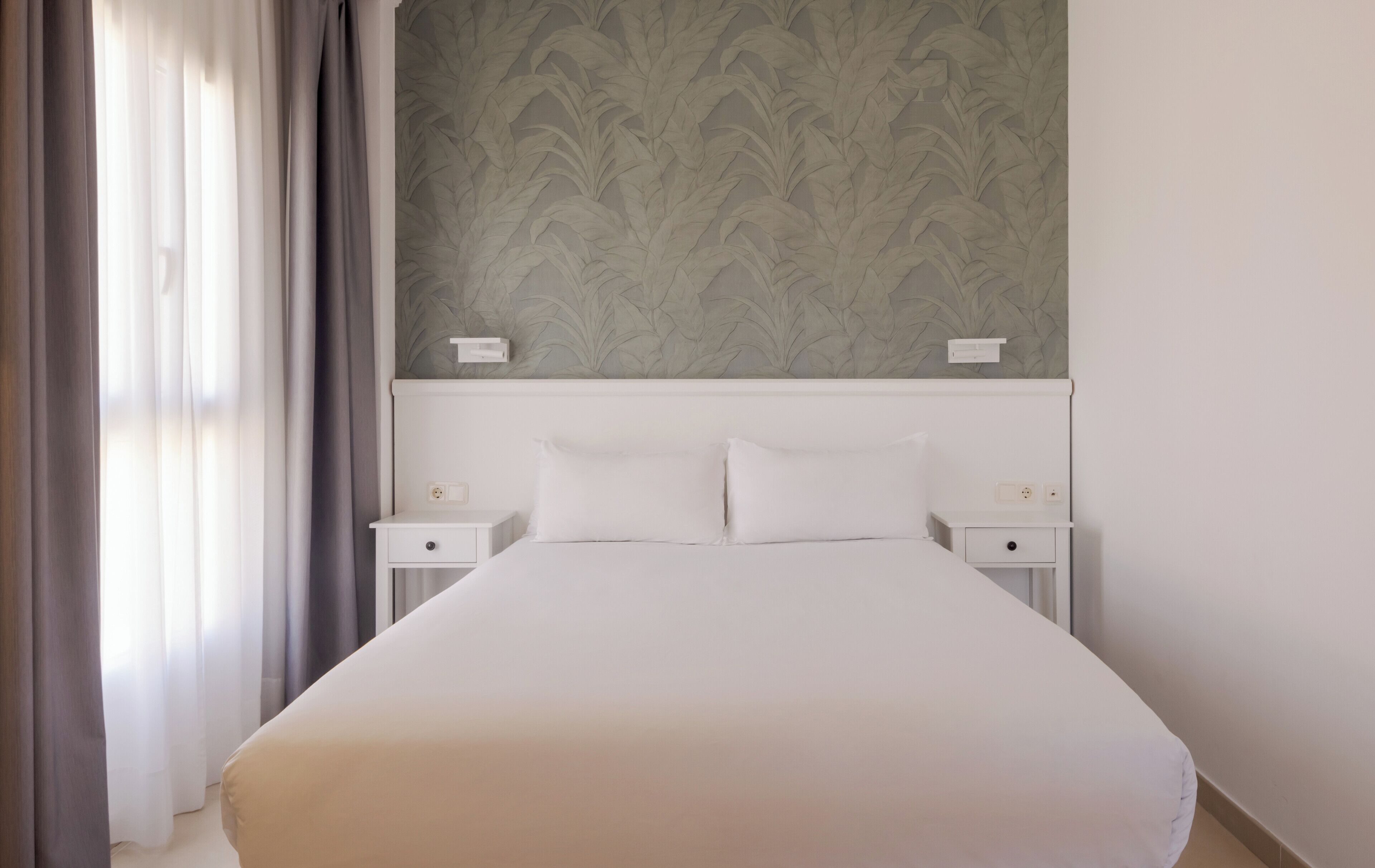 Quarto de hotel com uma cama grande, duas mesas de cabeceira, cabeceira com padrão de folha e uma janela com cortinas.