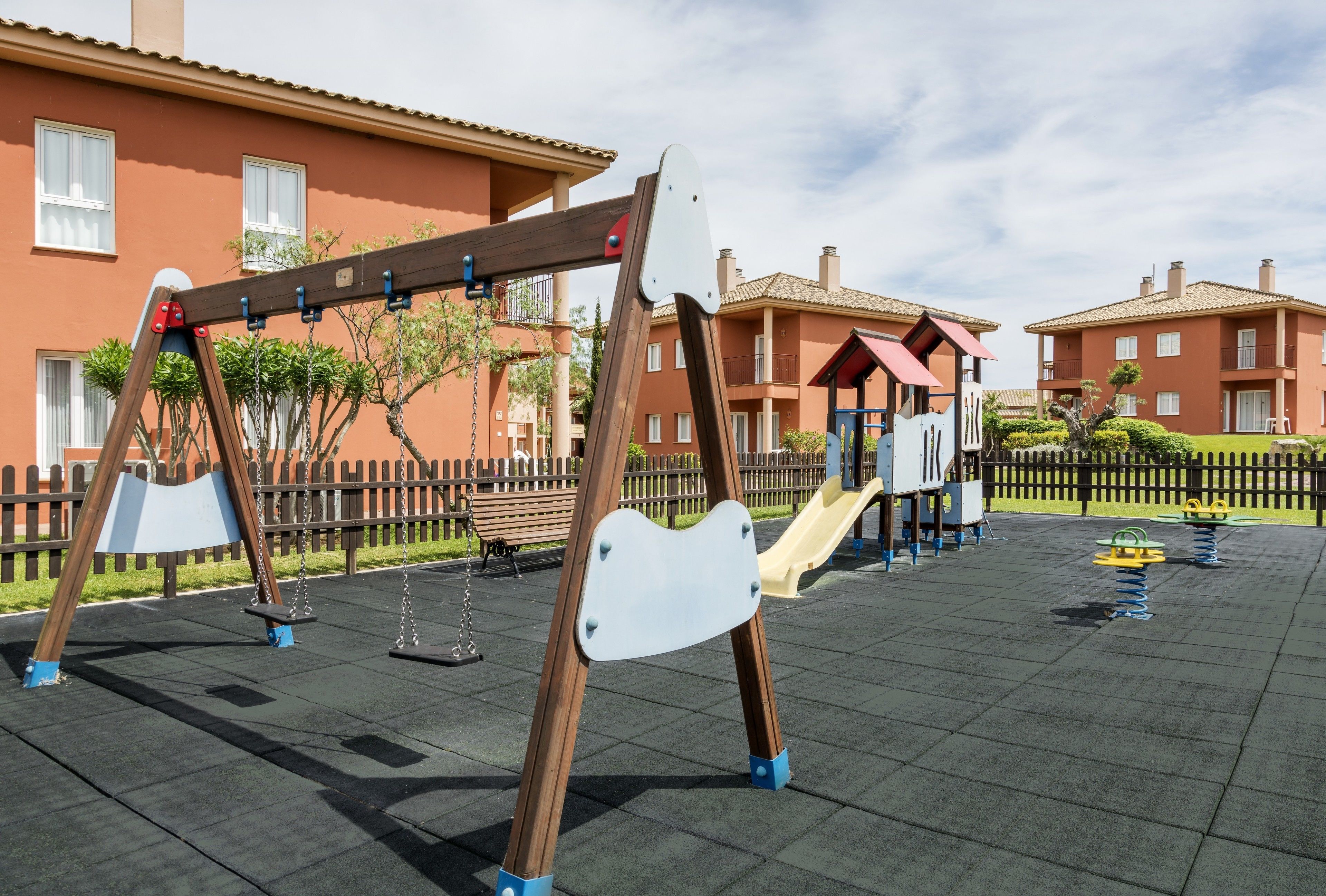 A playground with swings and a slide, surrounded by a fence and residential buildings.
