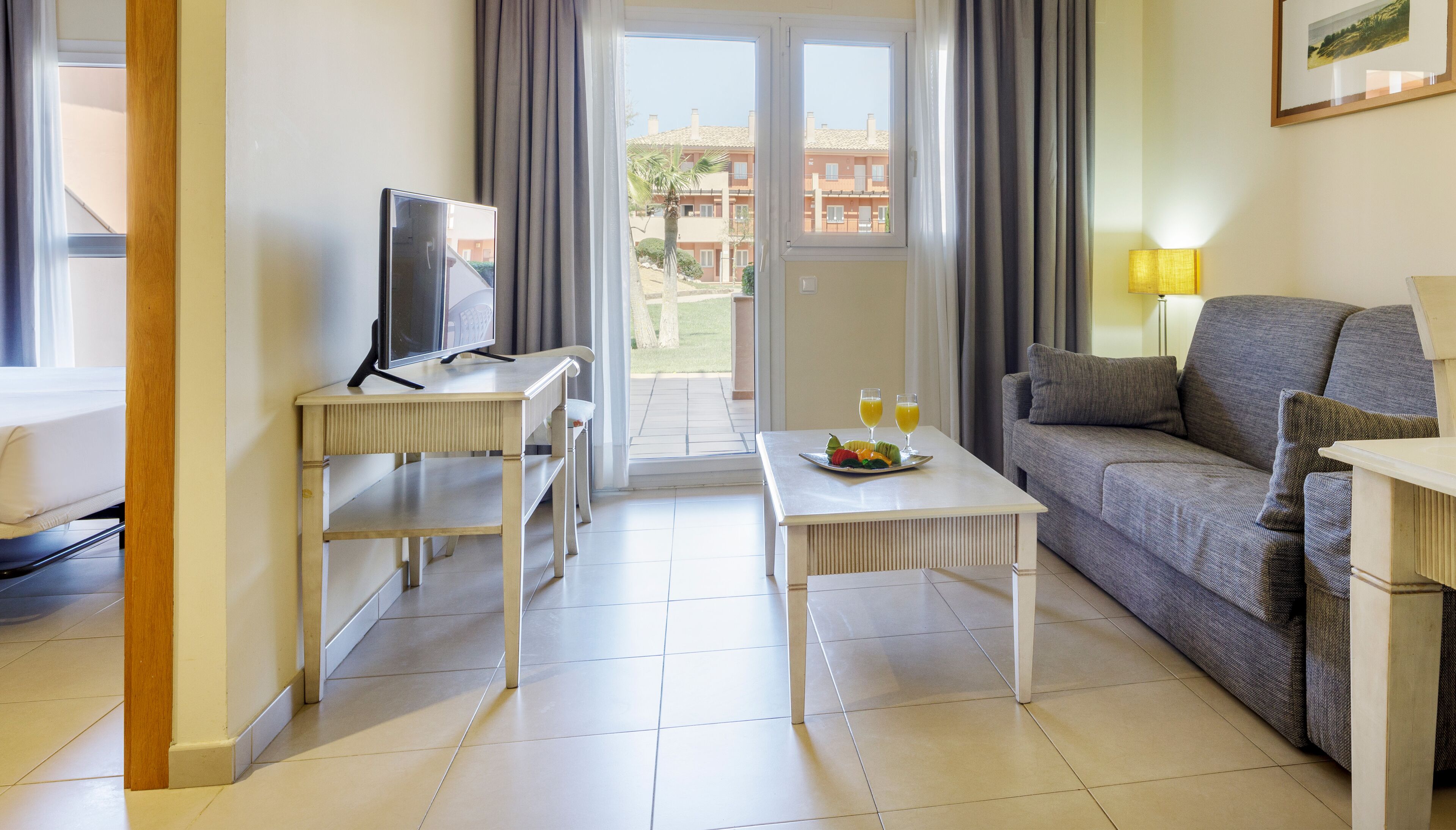 A hotel room with a bed, a TV on a desk, a sofa, a dining table with fruit and drinks, and a view of buildings outside.