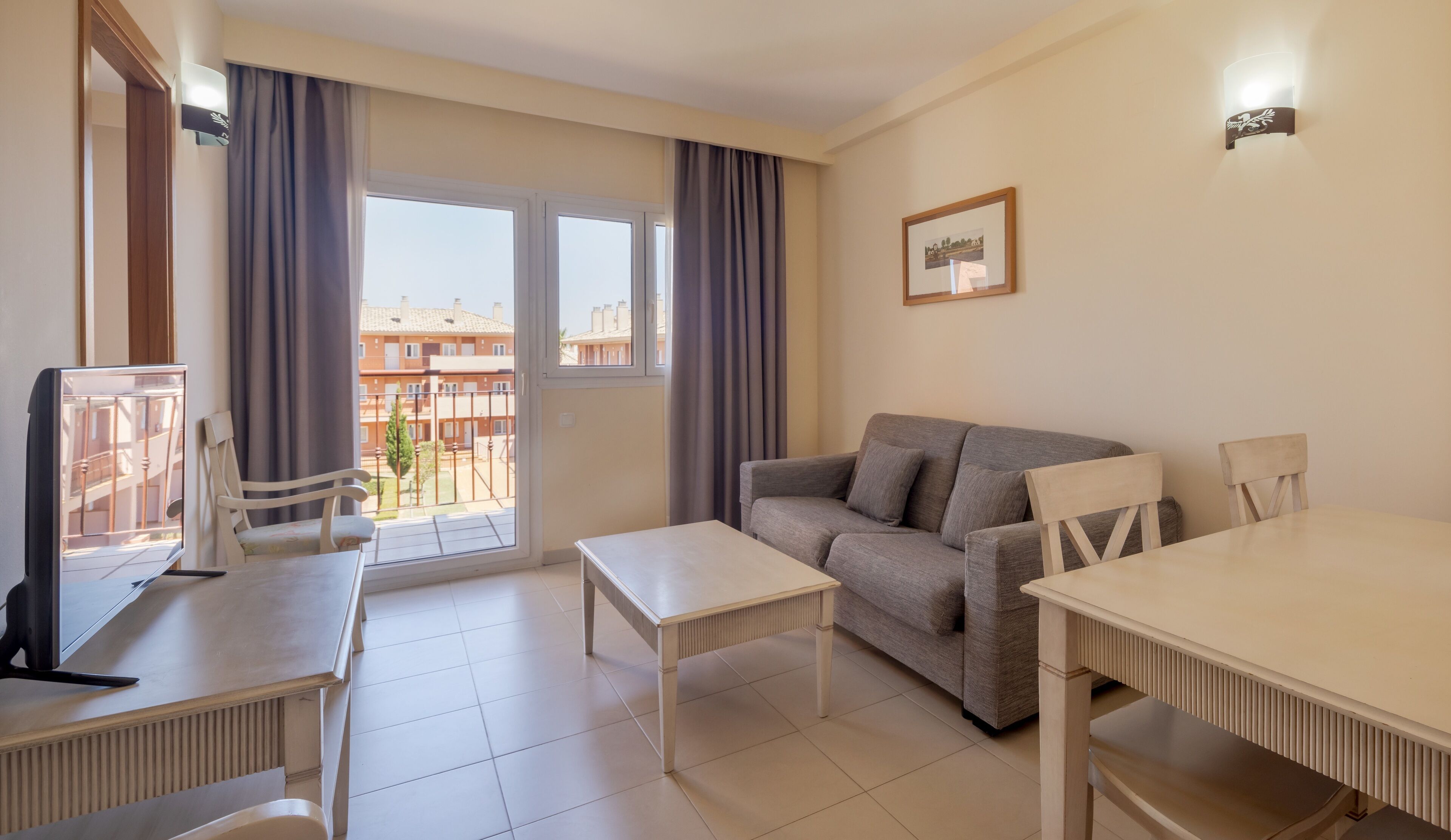 A living room with a sofa, a table, a TV, and a balcony with curtains.