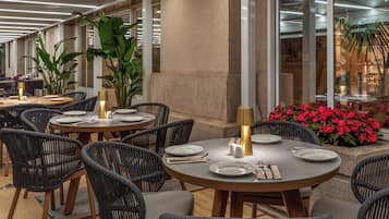 Un restaurante con mesas redondas, sillas de mimbre y plantas en macetas.