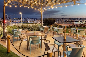 An outdoor seating area with tables and chairs, a pergola with string lights, and a cityscape view in the background.