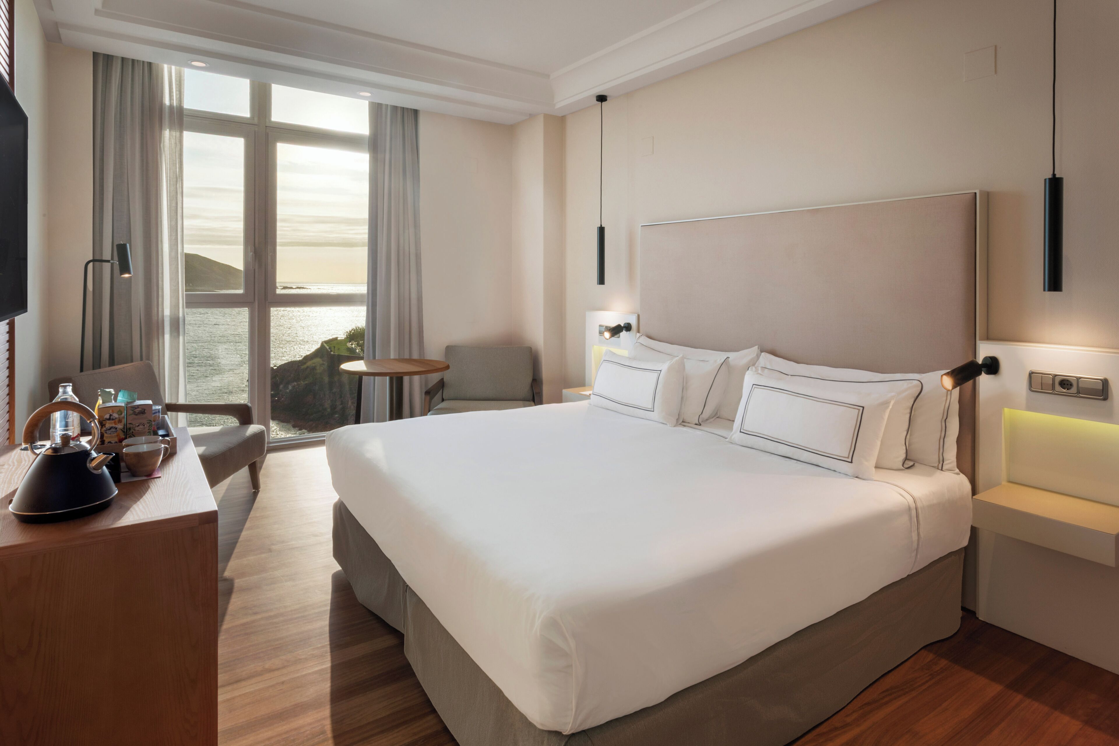 A hotel room with a large bed, a desk with a kettle, a chair, a small table, and a view of the sea.