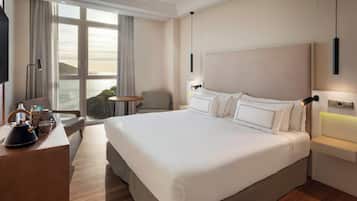 A hotel room with a large bed, a desk with a kettle, a chair, a small table, and a view of the sea.