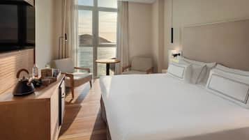 Habitación de hotel con cama, escritorio con hervidor, silla, mesita y ventana grande con vista al mar.