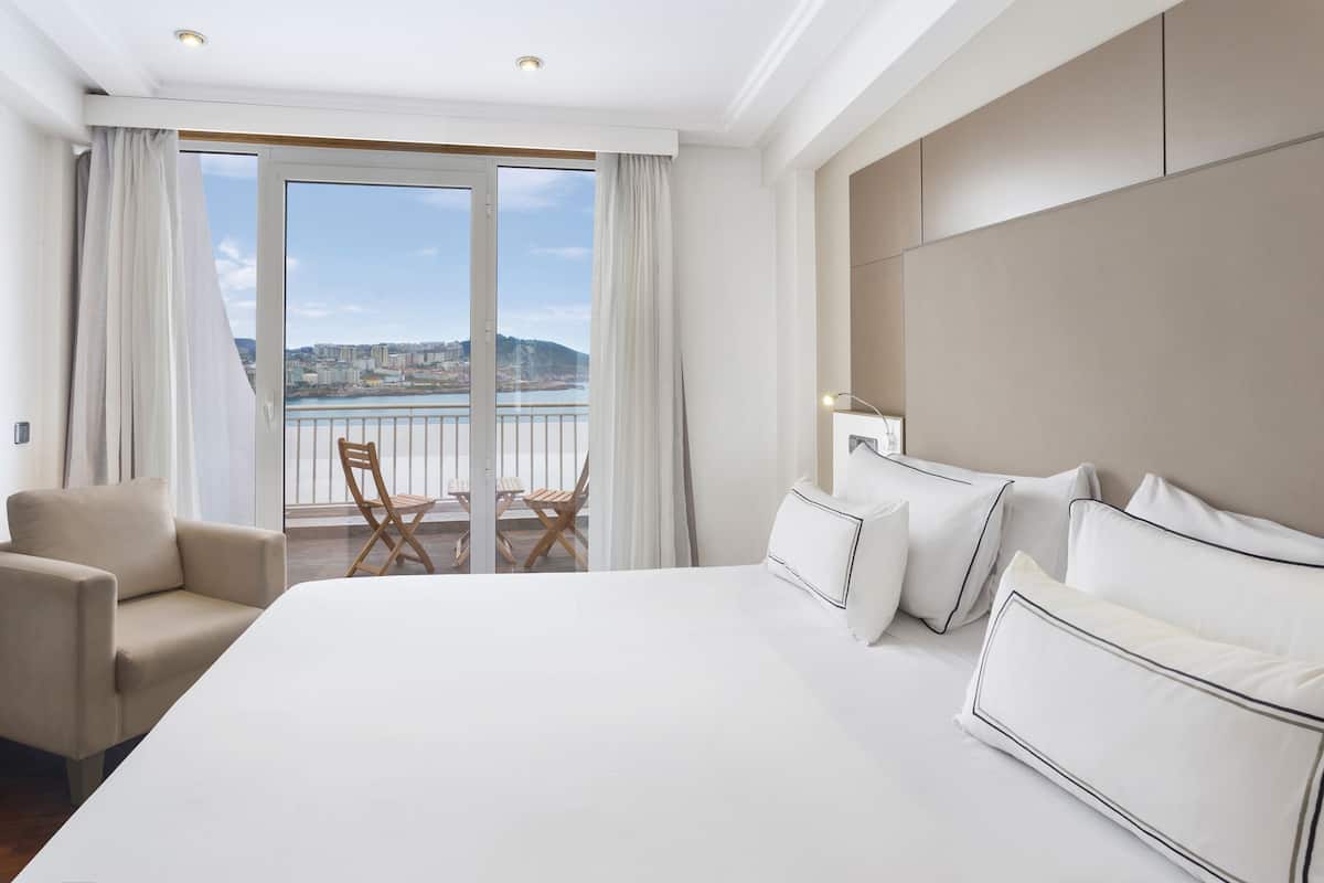 Ein Hotelzimmer mit einem großen Bett, einem Sessel, einem Balkon mit Blick auf die Stadt und einer durch das Fenster sichtbaren Stadtansicht.