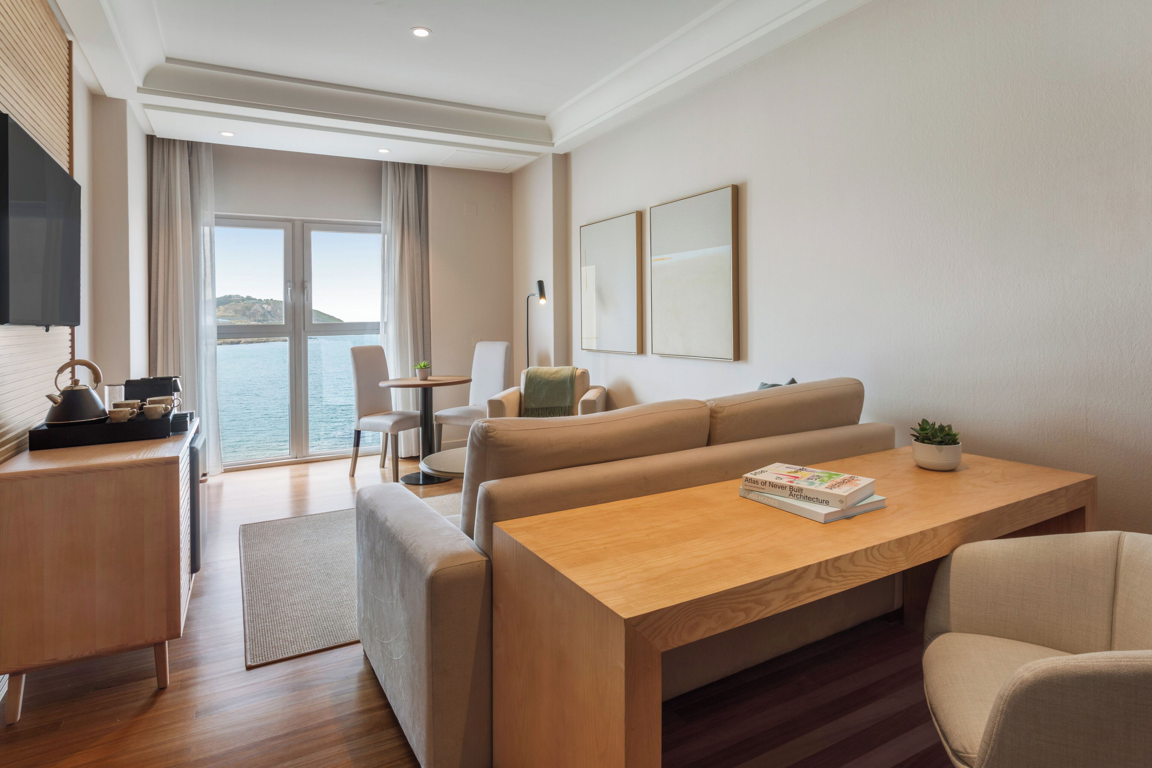 Een moderne hotelkamer met een bank, een houten bijzettafeltje, een televisie en uitzicht op zee.