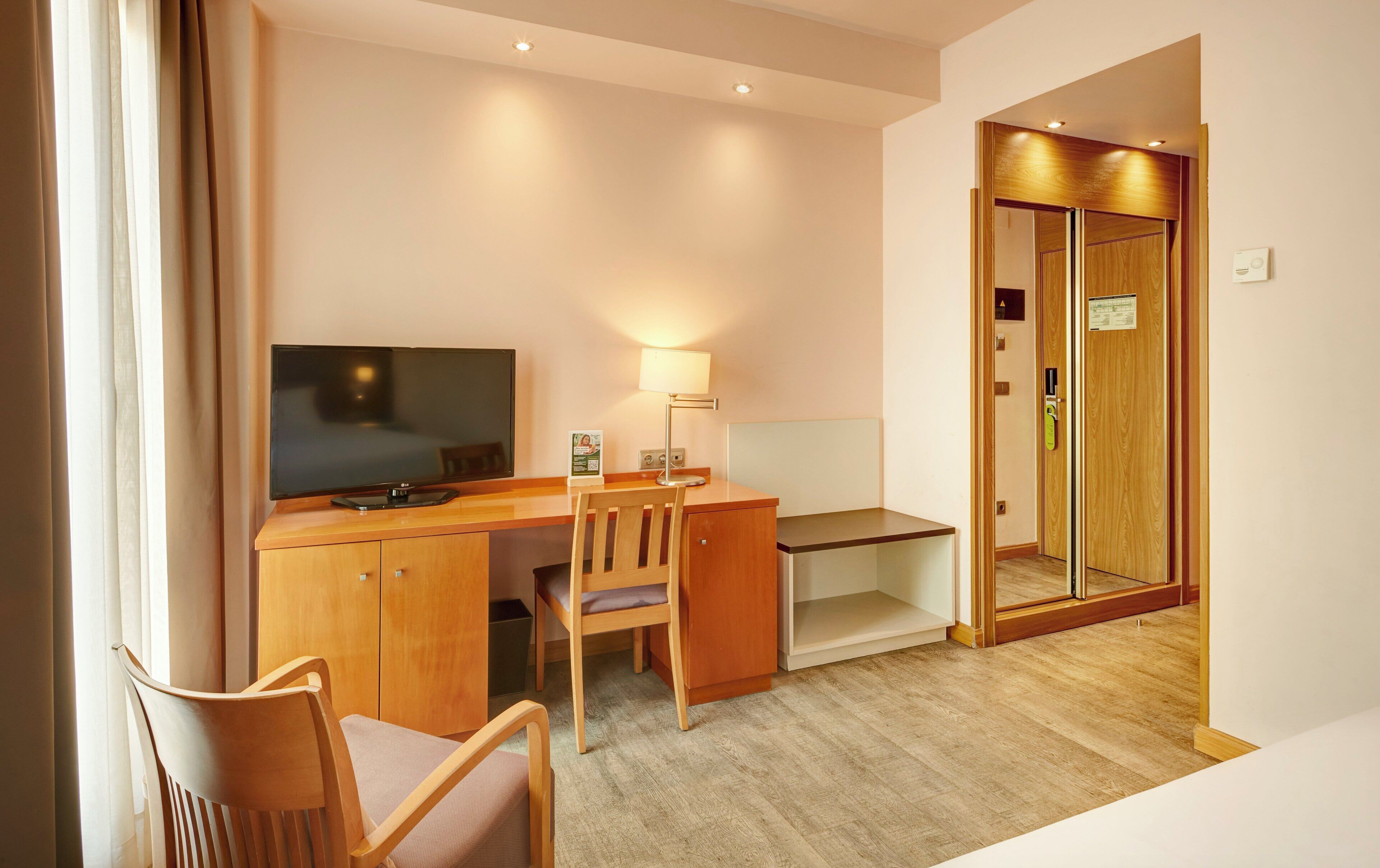Una camera d'albergo con una scrivania in legno, una sedia, un televisore, una lampada e una porta che dà su un'altra stanza.