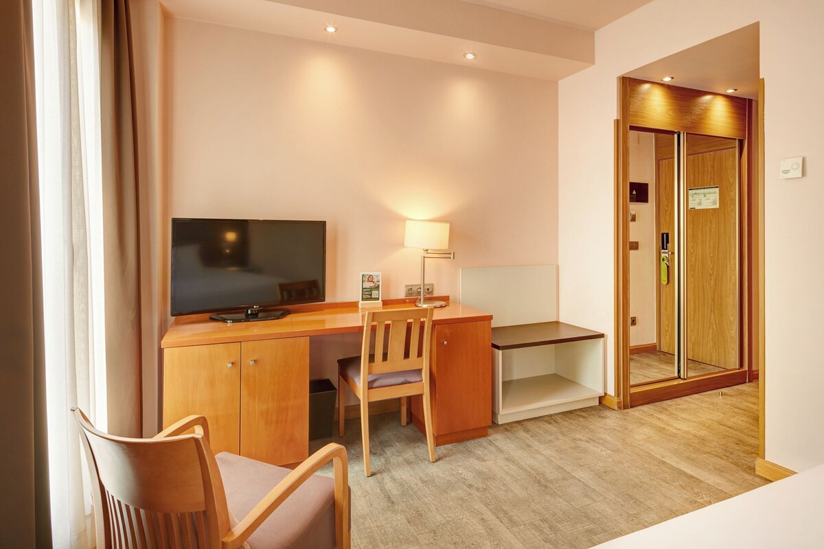 A hotel room with a wooden desk, a chair, a television, a lamp, and a mirror.