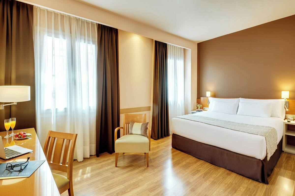 Quarto de hotel com uma cama, uma mesa com um laptop, uma cadeira e uma mesa com um copo de suco de laranja.